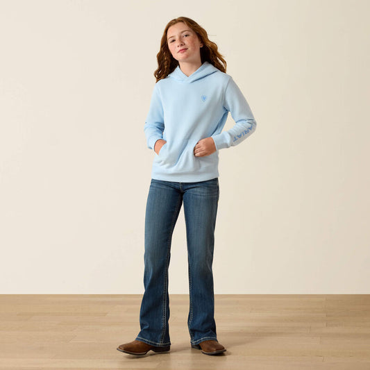 Person wearing a light blue hoodie and jeans standing on a wooden floor with a beige wall background