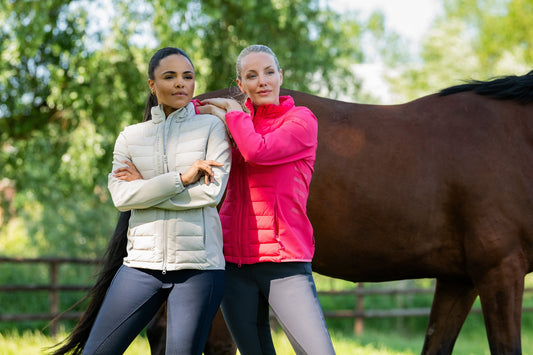 Two girls in jackets standing next to a horse in a grassy setting