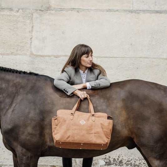 A person standing beside a chestnut-colored horse, holding a chestnut weekend travel bag with a zipper.